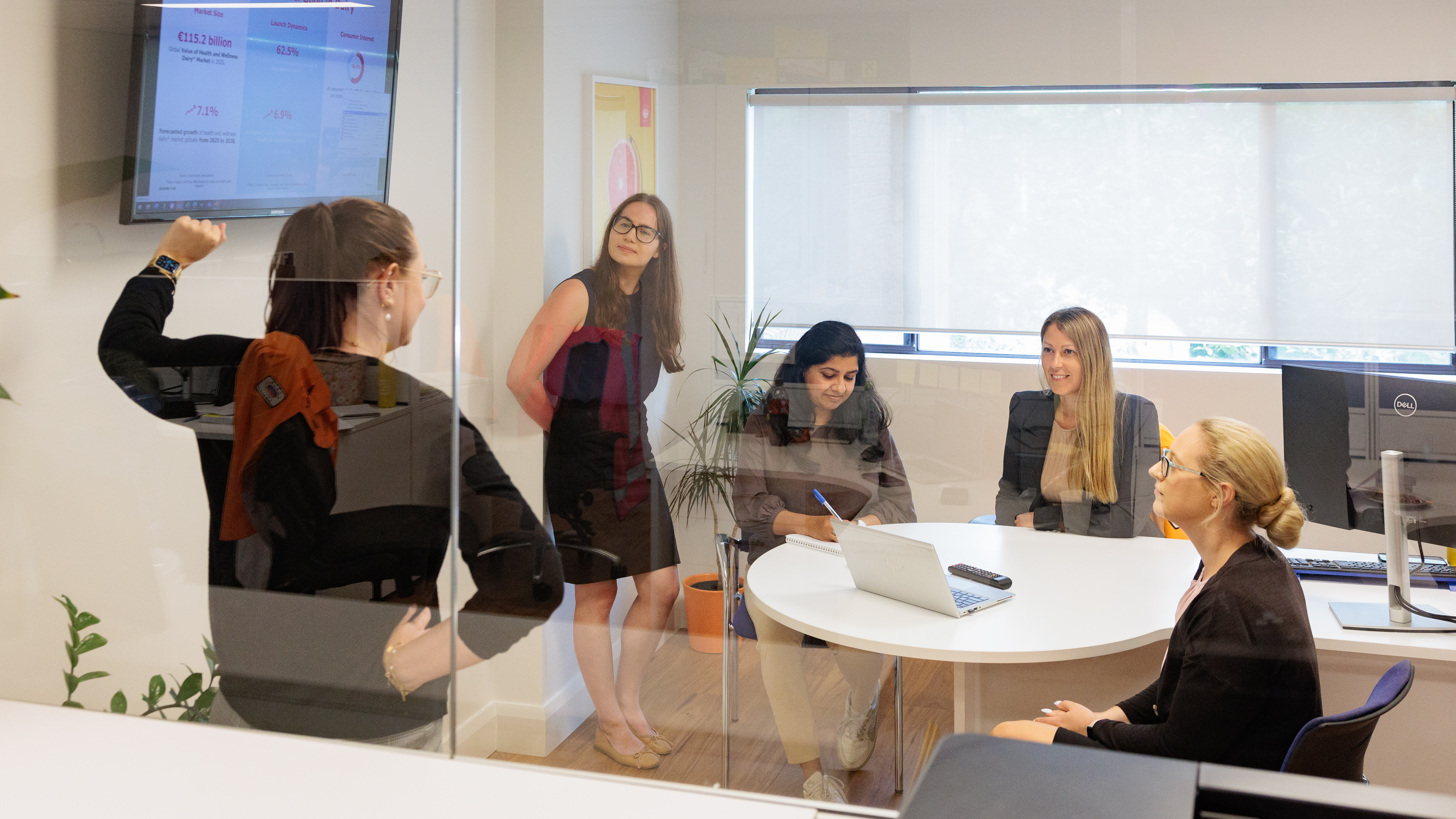 Five colleagues in a glass-walled meeting room during a presentation