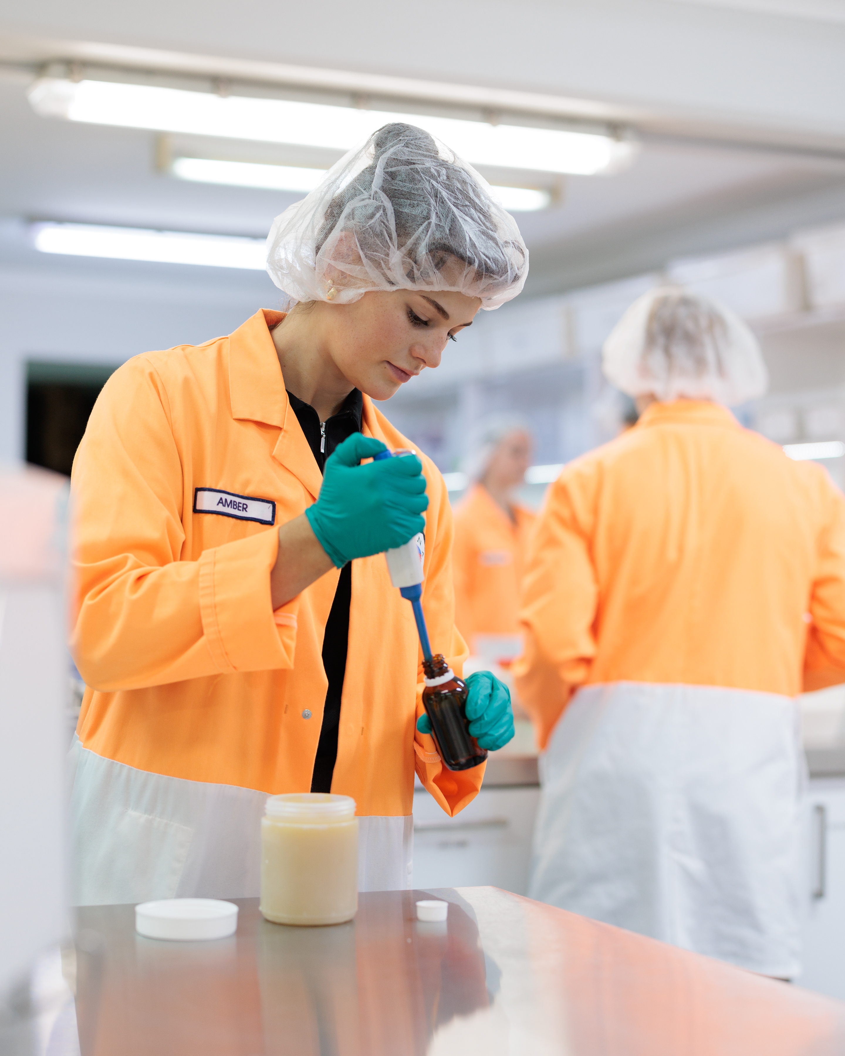 Lab employee in an orange coat and hairnet pipetting a sample into a jar at the Australian plant