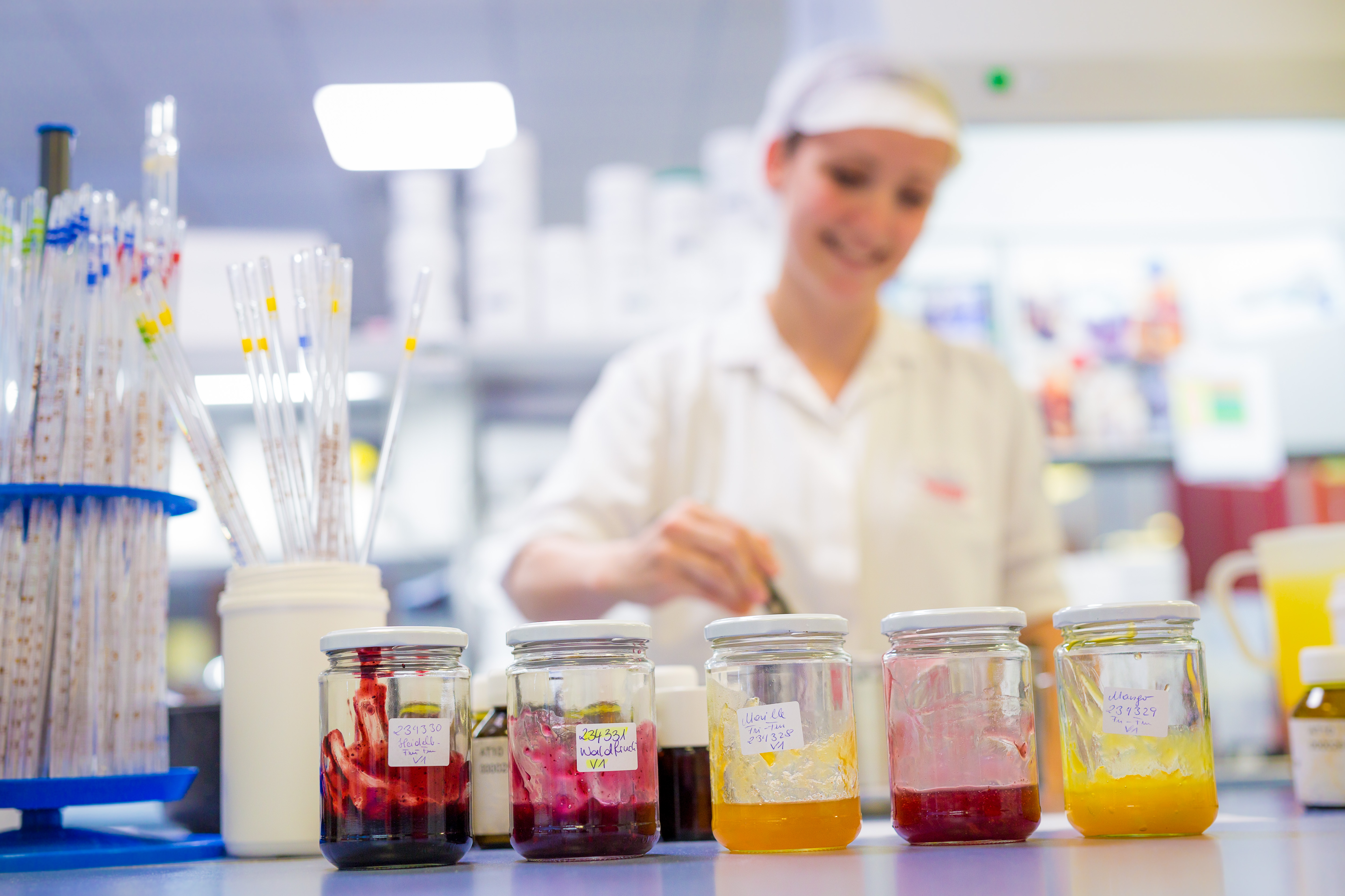 Row of labelled jars with fruit preparation samples on a lab bench, a technician in white working behind them [no AI]