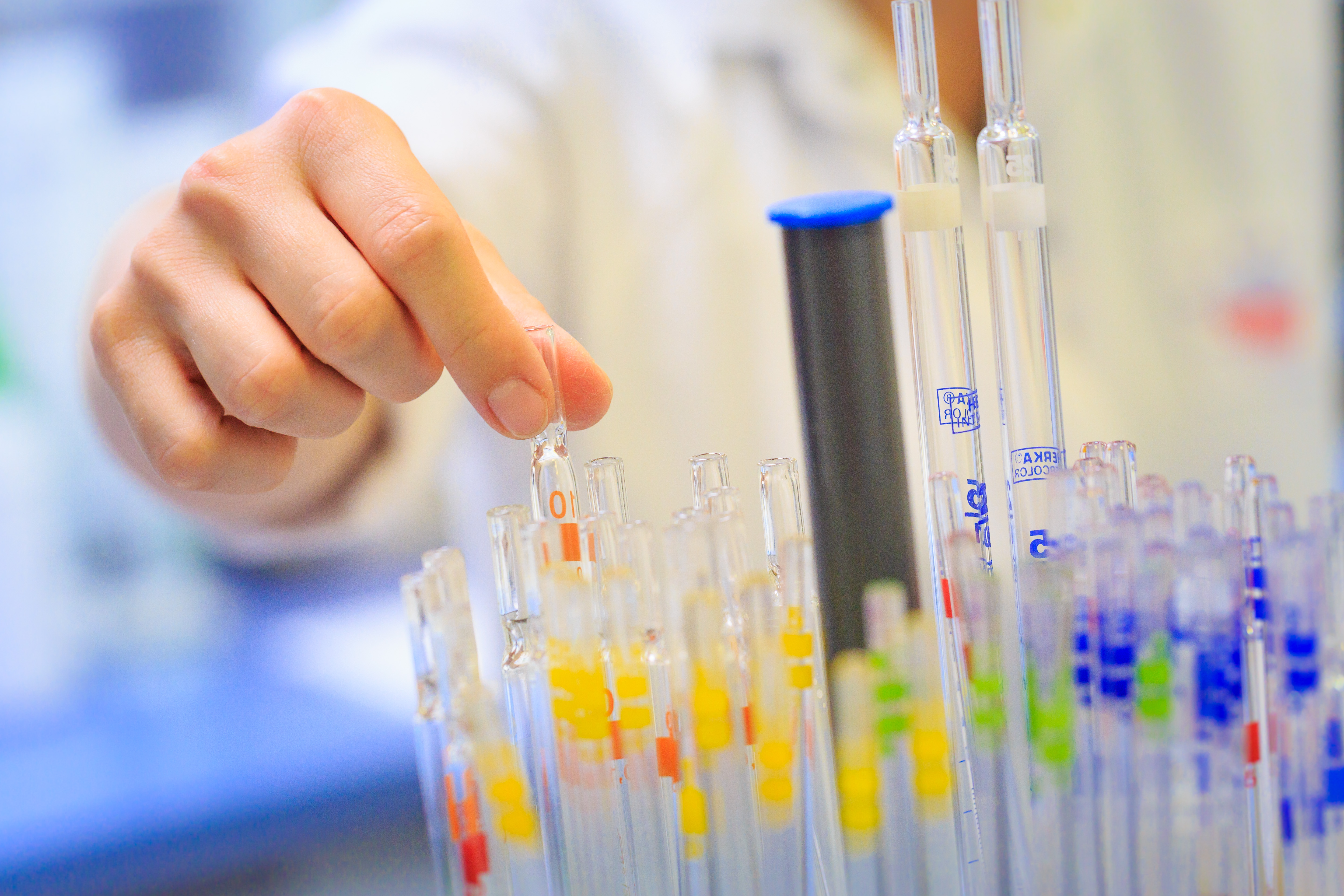 Hand reaching for a glass pipette in a laboratory rack of colour-coded pipettes [no AI]