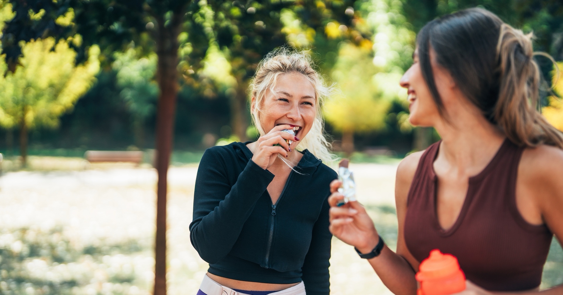 Two women in sportswear laughing and eating a snack bar in a park [no AI]