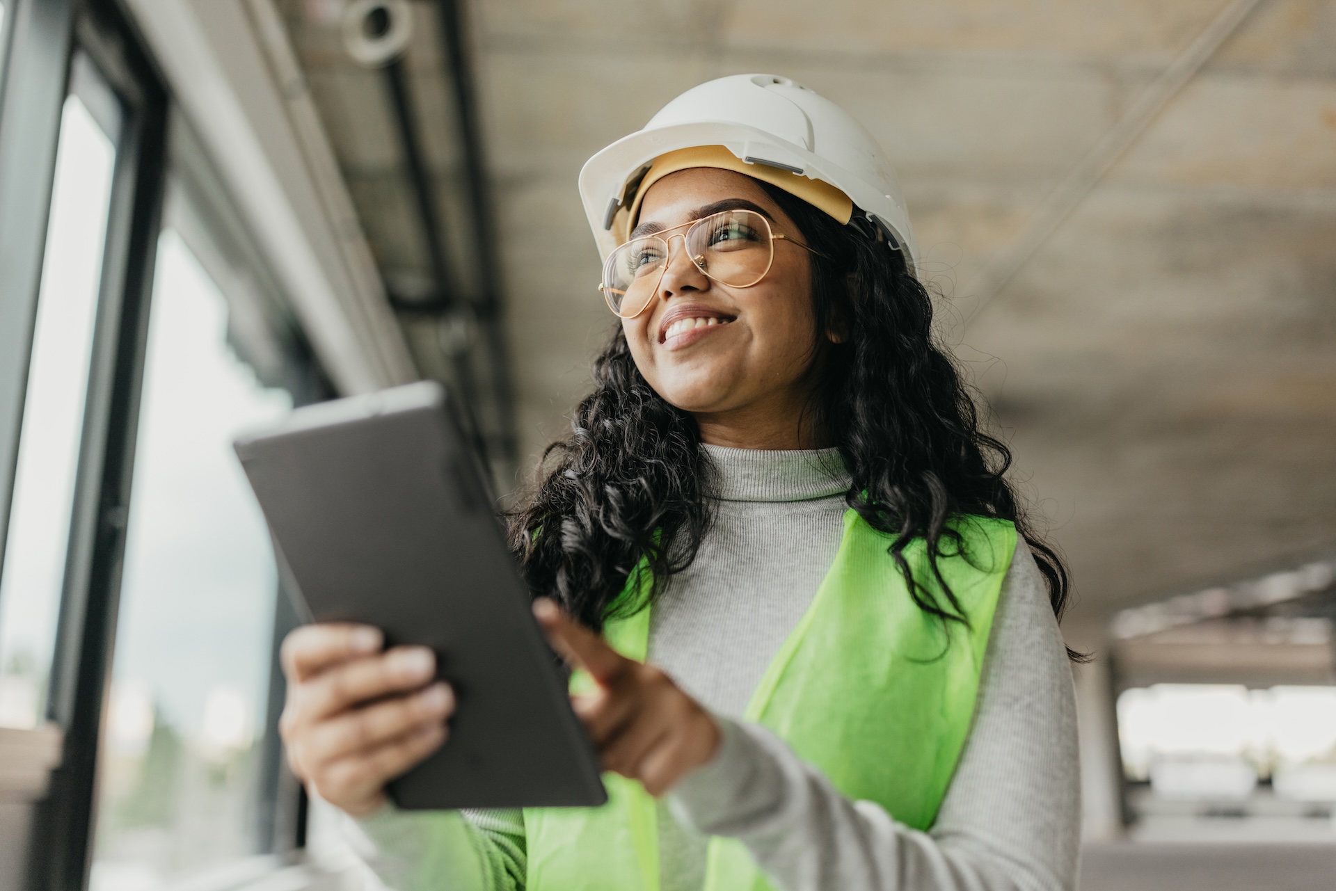 Smiling engineer in a white hard hat and safety vest holding a tablet at an industrial site [no AI]