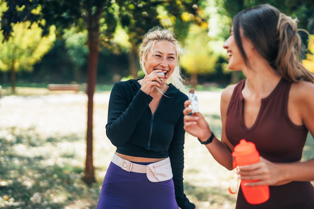 Zwei Frauen in Sportkleidung lachen in einem sonnigen Park, eine mit Trinkflasche, eine trinkt aus einem Fläschchen [no AI]
