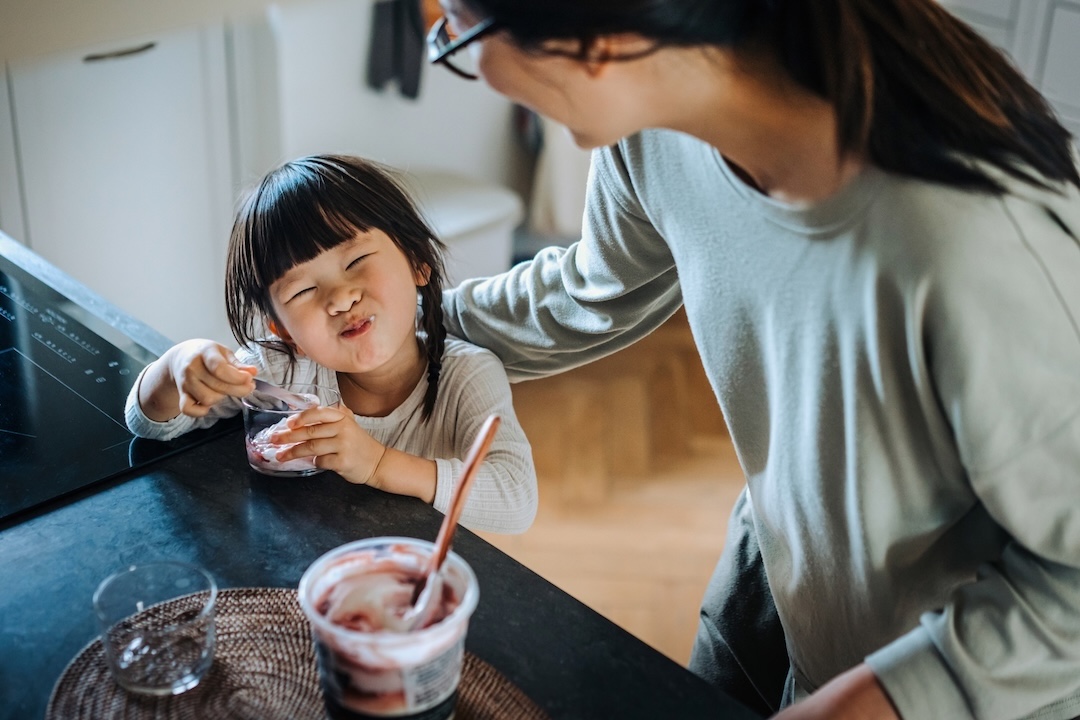 Mother and grinning daughter eating a dessert from a cup together at the kitchen counter [no AI]