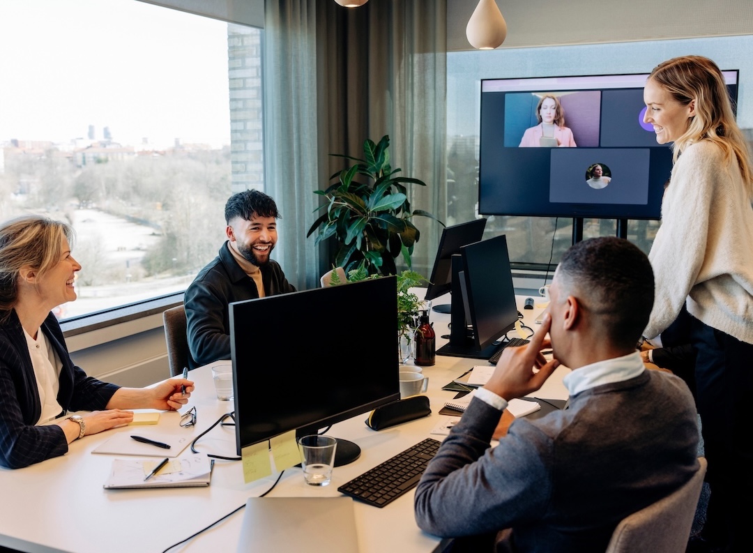 Colleagues in an office talking and smiling, a video call on the screen behind them [no AI]