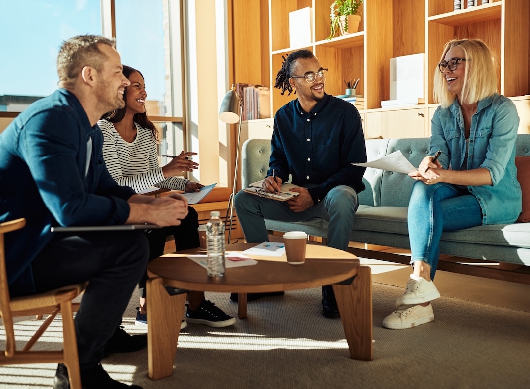 Four colleagues laughing together during a relaxed meeting in a sunlit lounge [no AI]