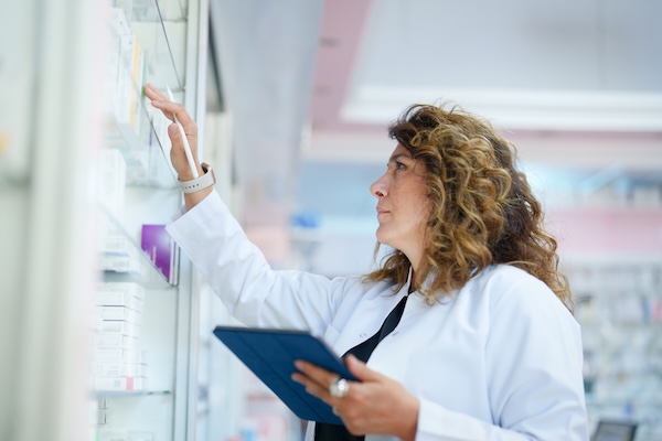 Apothekerin im weißen Kittel mit Tablet prüft Produkte in einem Regal [no AI]
