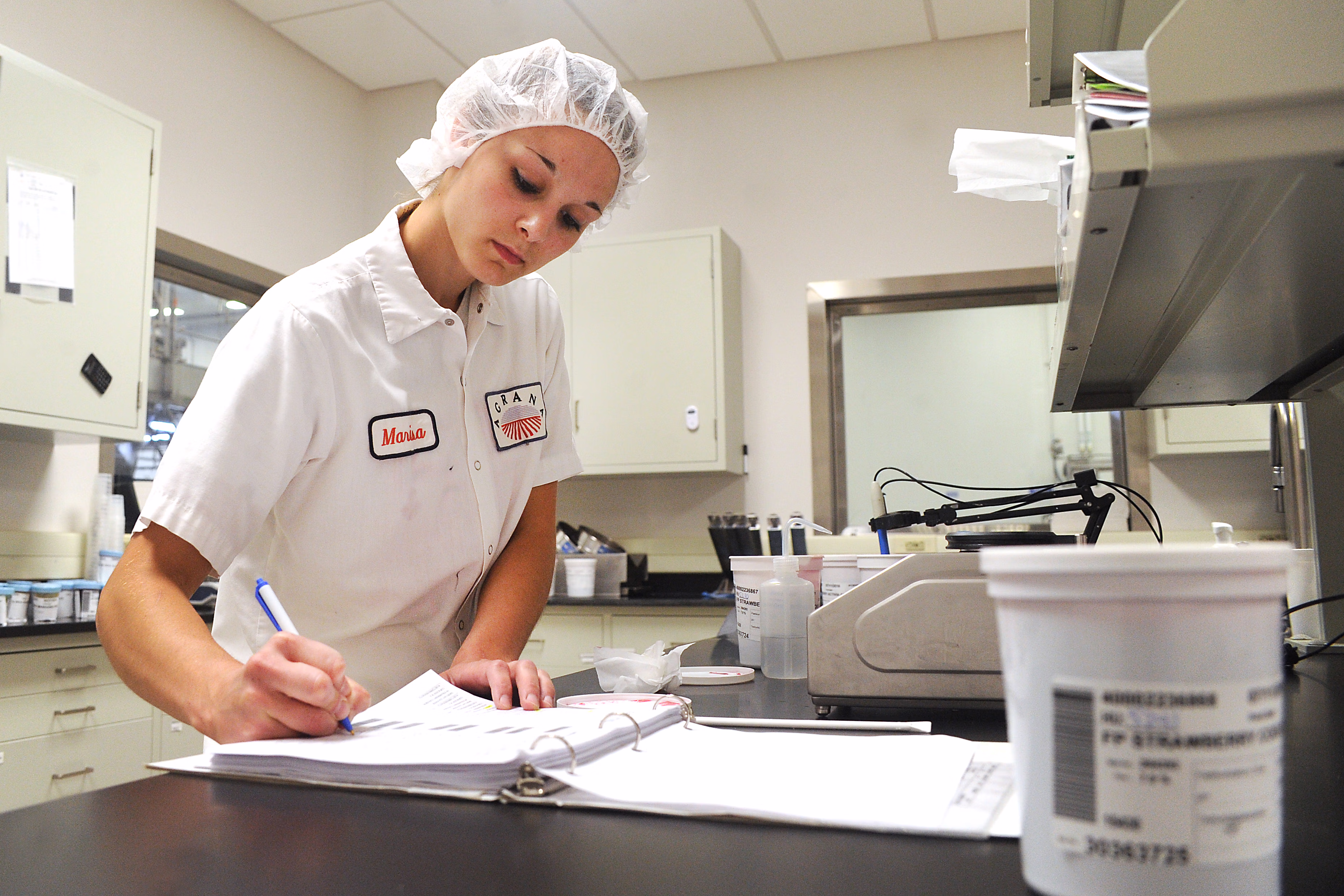 Lab technician in hairnet making notes in a binder in the quality lab of a US plant [no AI]
