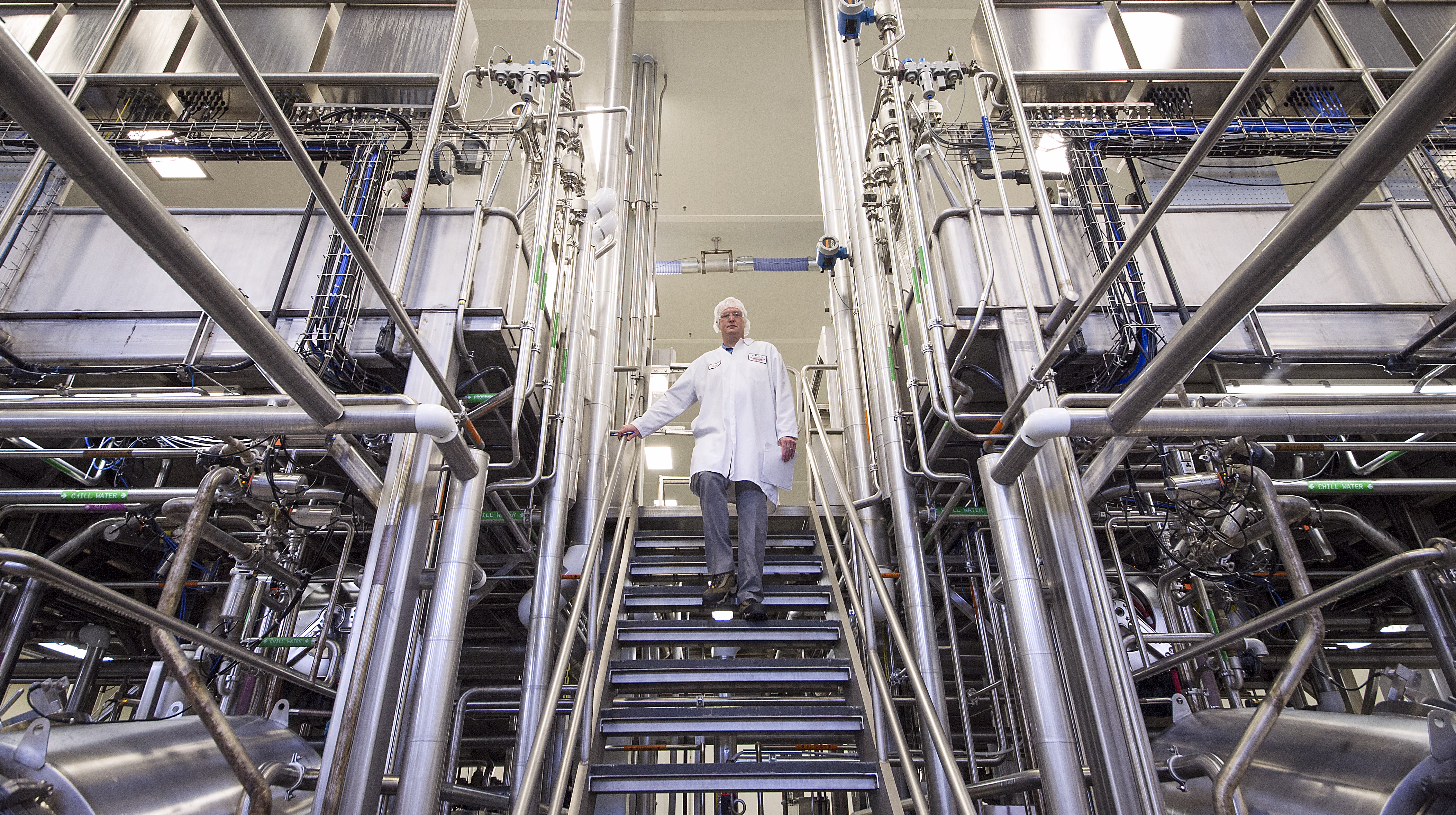 Employee in a white coat walking down stairs between stainless steel process pipes in a US plant [no AI]
