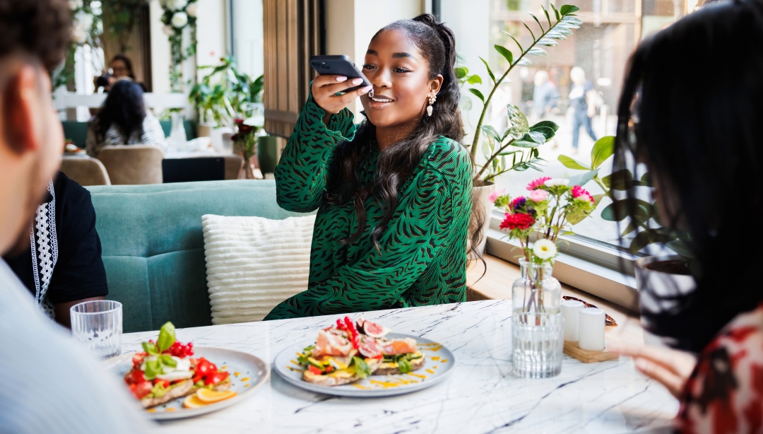 Woman in a green dress photographing her plate with a smartphone at a restaurant table with friends [no AI]