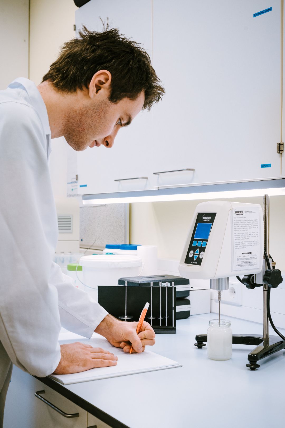Young man in a lab coat taking notes next to a viscometer with a sample in a laboratory