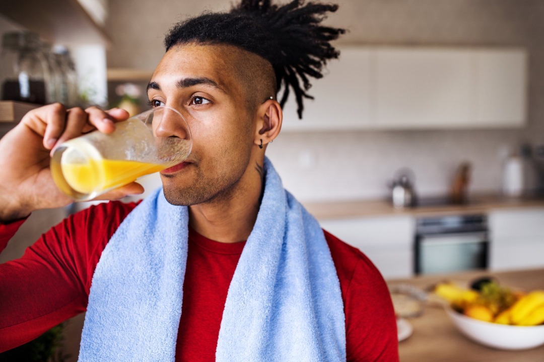 Junger Mann mit Handtuch um den Hals trinkt ein Glas Orangensaft in einer Küche [no AI]