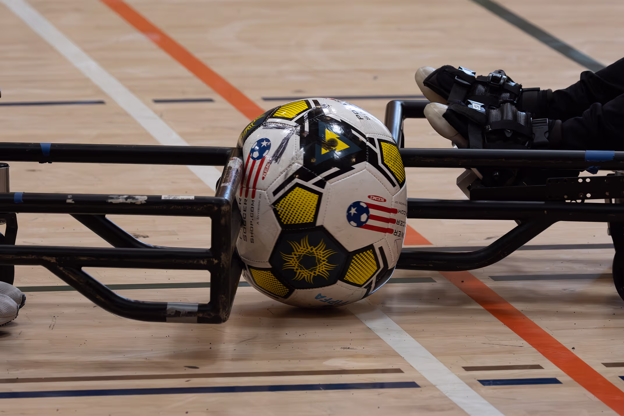 Close-up of a yellow, black, and white soccer ball held by a metal frame in a gymnasium with a wooden floor marked with sport lines.