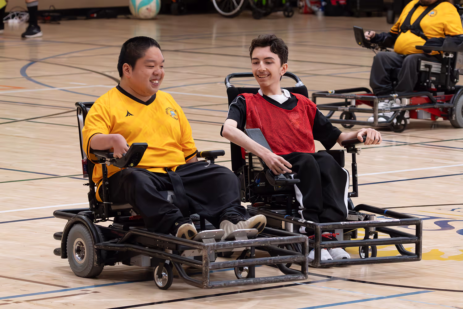 Two young men in powered wheelchairs wearing sports jerseys smiling together on an indoor gym court.