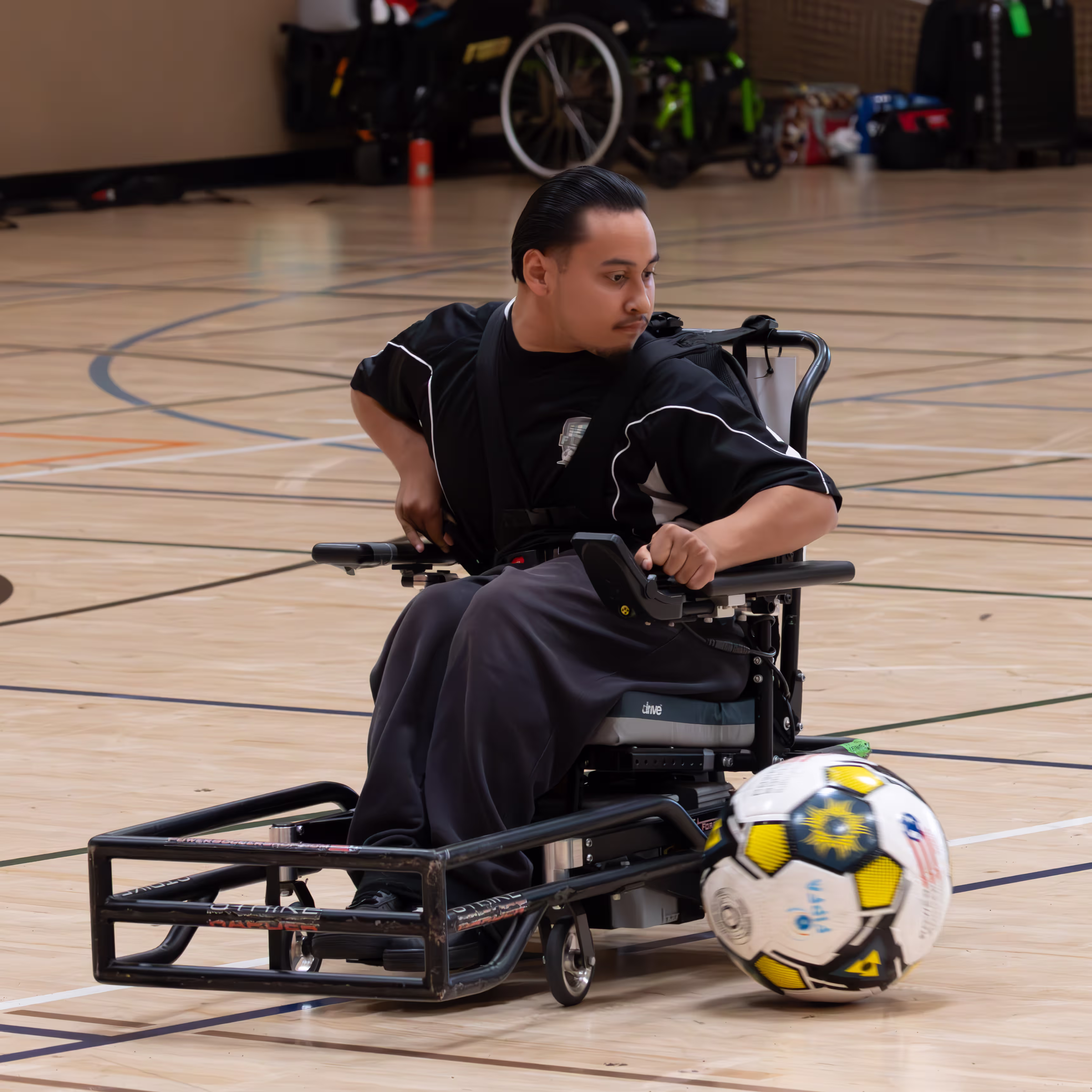 Man in a motorized wheelchair playing power soccer indoors on a gym floor, controlling a large soccer ball with a front bumper.
