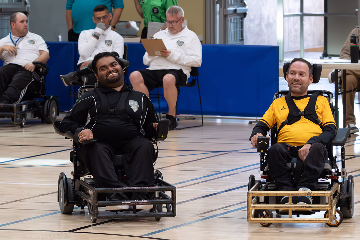 Two men in power wheelchairs smiling on a gym floor during a wheelchair soccer event, with others observing in the background.