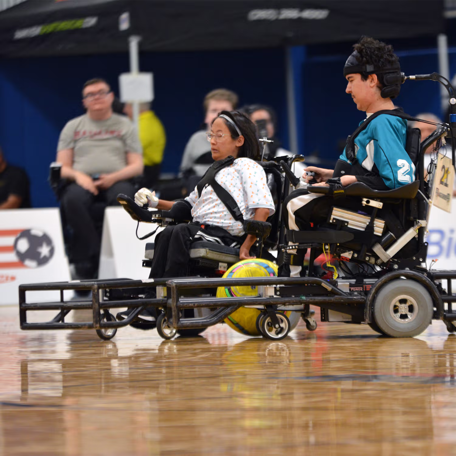 Two athletes in power wheelchairs playing power soccer indoors with a yellow ball between them.