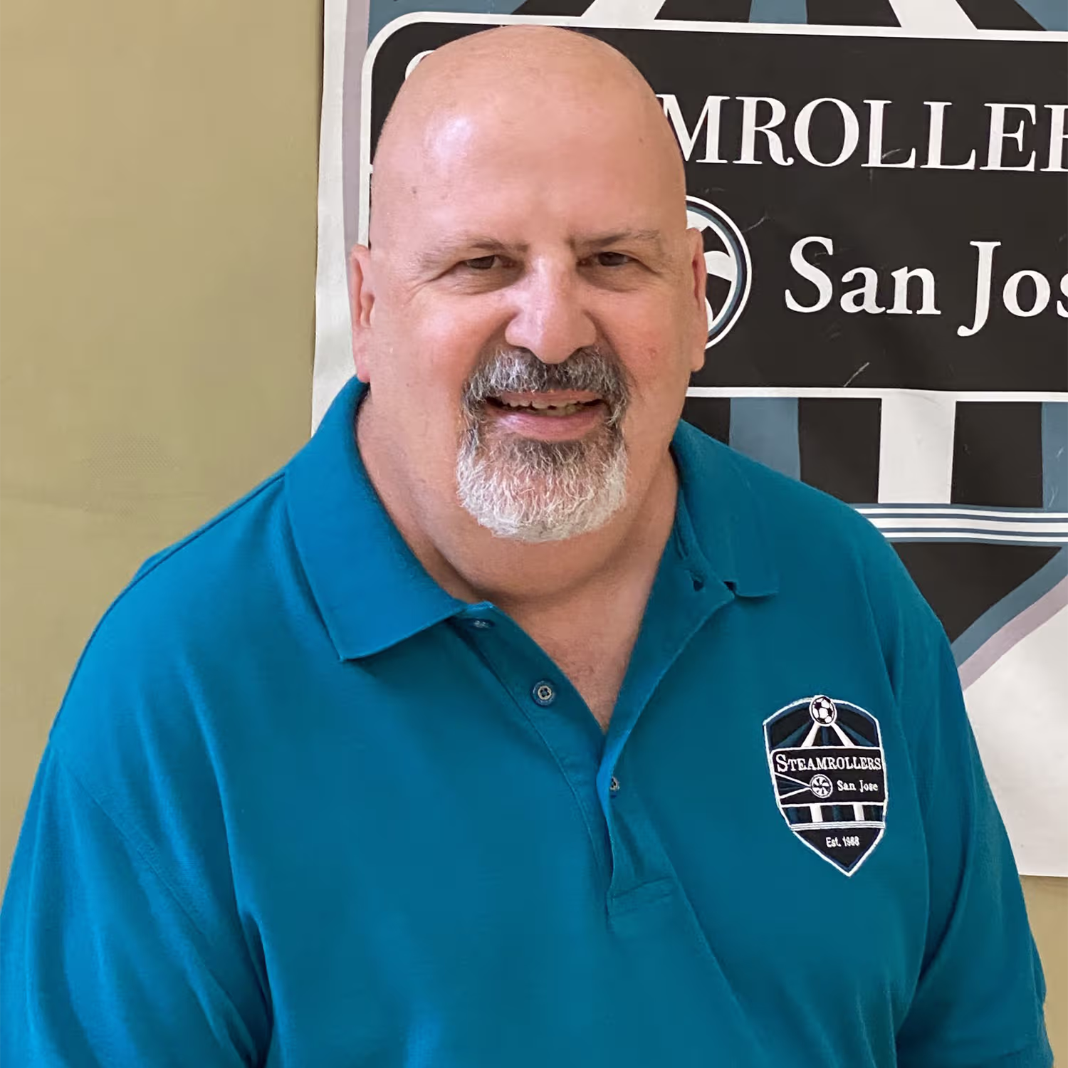 Smiling bald man with a gray goatee wearing a teal polo shirt with a San Jose Steamrollers logo.