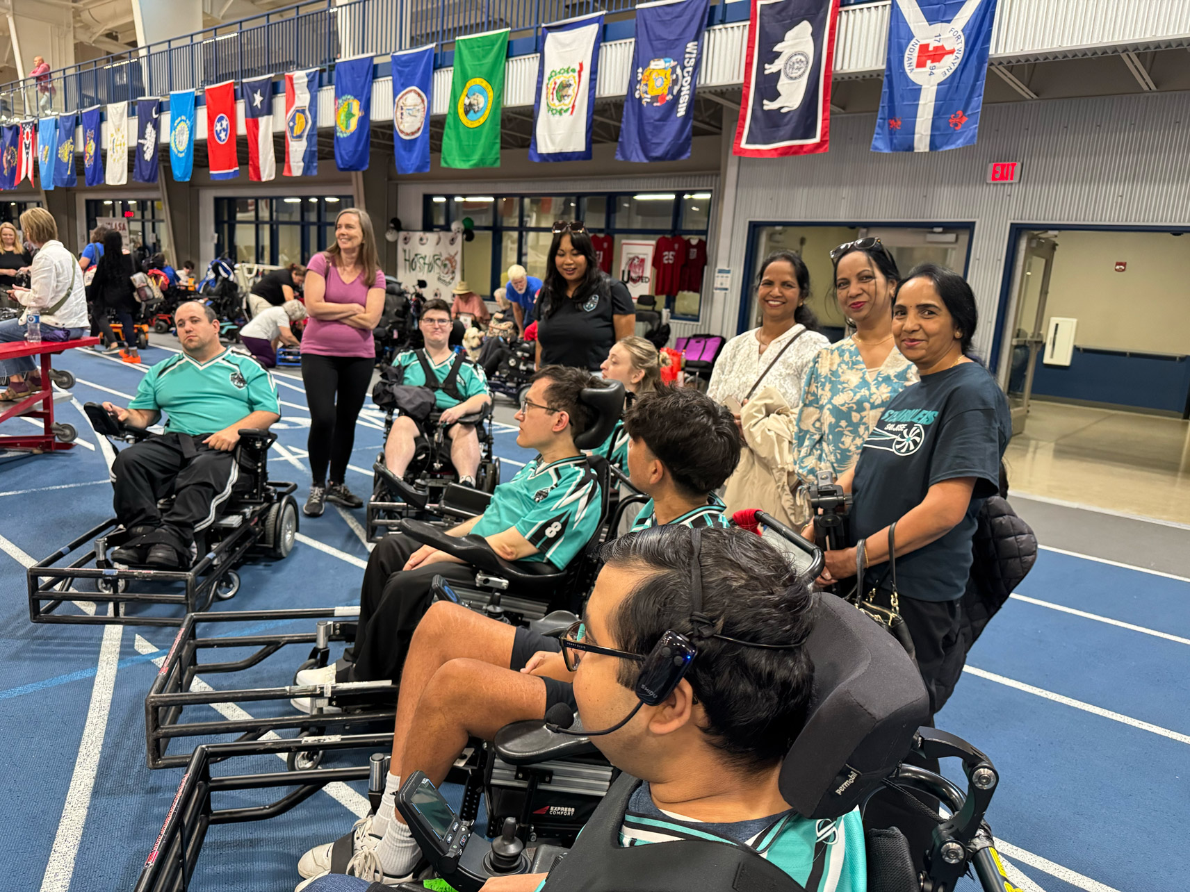 Group of athletes in power wheelchairs wearing teal jerseys on an indoor track, accompanied by standing supporters, with various state flags hanging above.