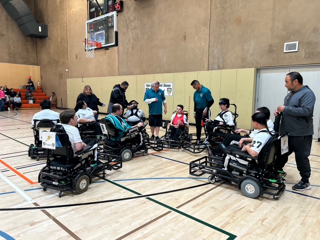 Group of athletes in power wheelchairs gathered in a circle on an indoor basketball court with coaches and spectators nearby.