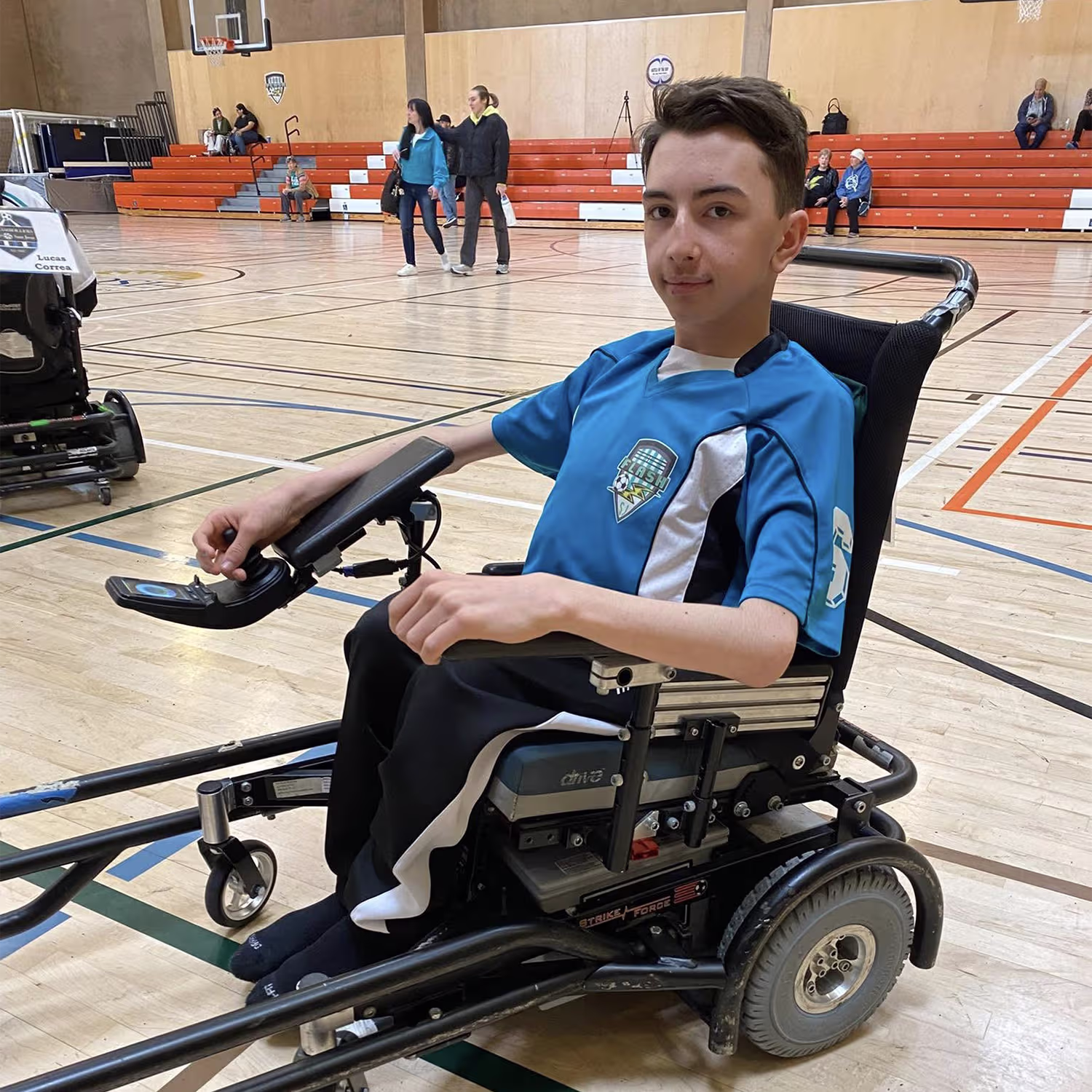 Teen boy in a blue sports jersey sitting in a powered wheelchair on a gymnasium basketball court.