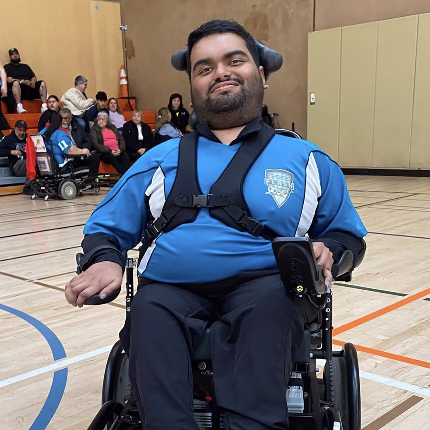 Man in a wheelchair wearing a blue sports jersey smiling in a gymnasium with spectators in the background.
