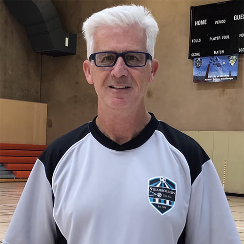 Man with white hair and glasses wearing a black and white Steamrollers jersey standing in an indoor basketball court.