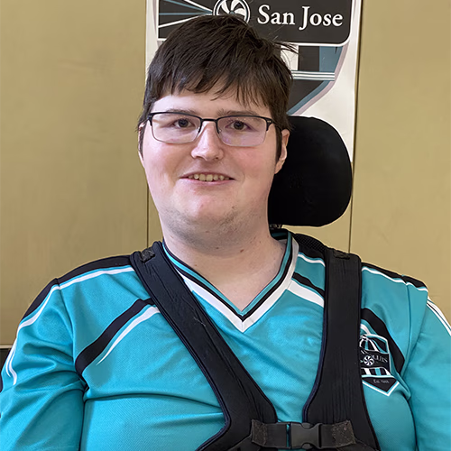 Smiling young person with short brown hair and glasses wearing a teal sports jersey seated in a wheelchair.