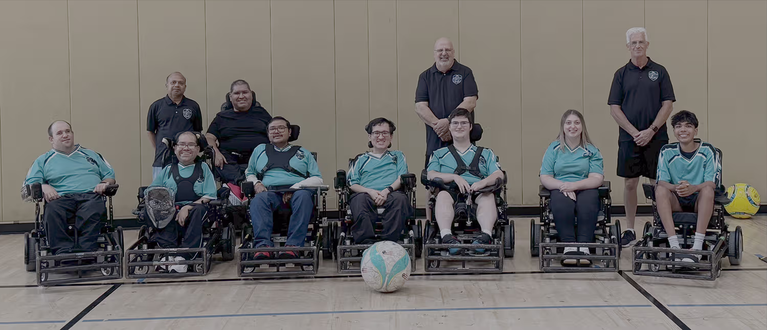 A group of eight wheelchair soccer players in teal jerseys with three standing coaches posing in a gym with a soccer ball in front.