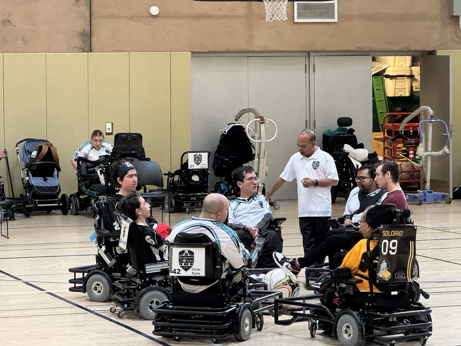 A group of seven wheelchair soccer players in power wheelchairs, gathered indoors on a gym court around a soccer ball, with a coach standing nearby talking to them.