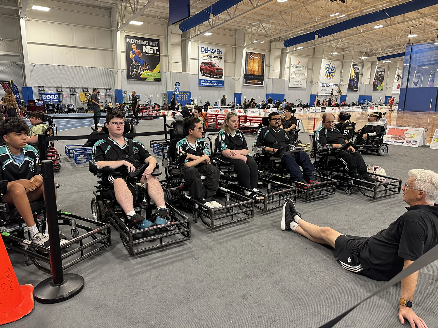 Group of athletes in power wheelchairs lined up indoors wearing matching black and teal uniforms, with a coach sitting on the floor in front of them.
