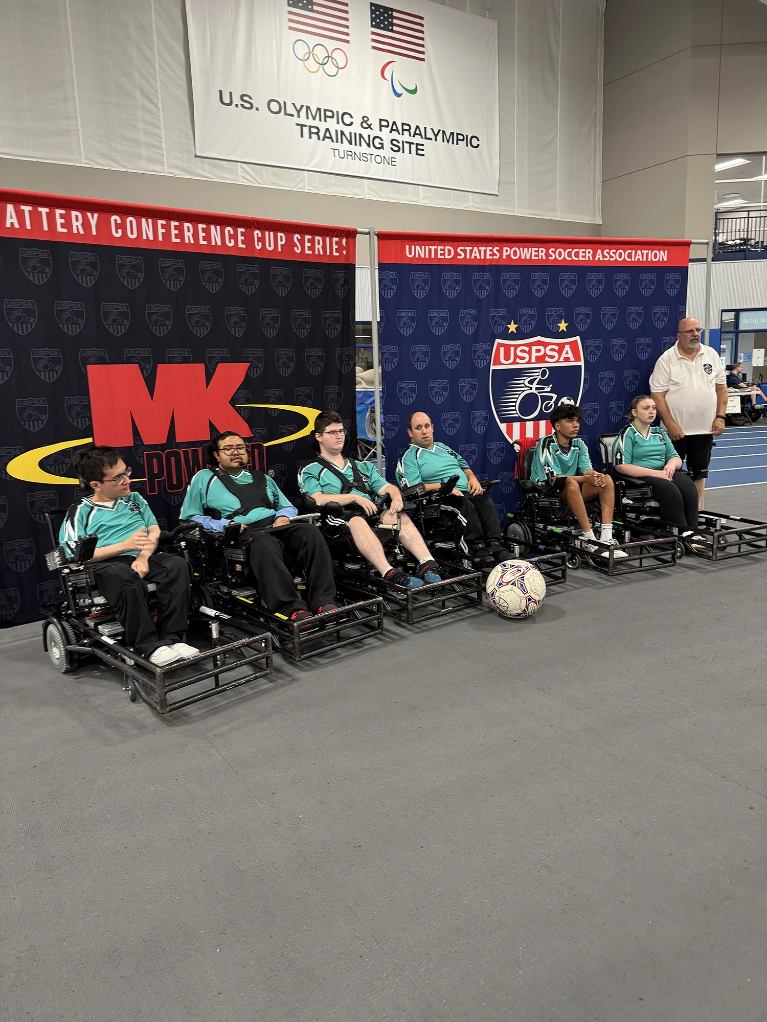 Six power soccer players in wheelchairs wearing teal jerseys sitting in a row indoors with a soccer ball in front and banners for U.S. Olympic & Paralympic Training Site and United States Power Soccer Association behind them.