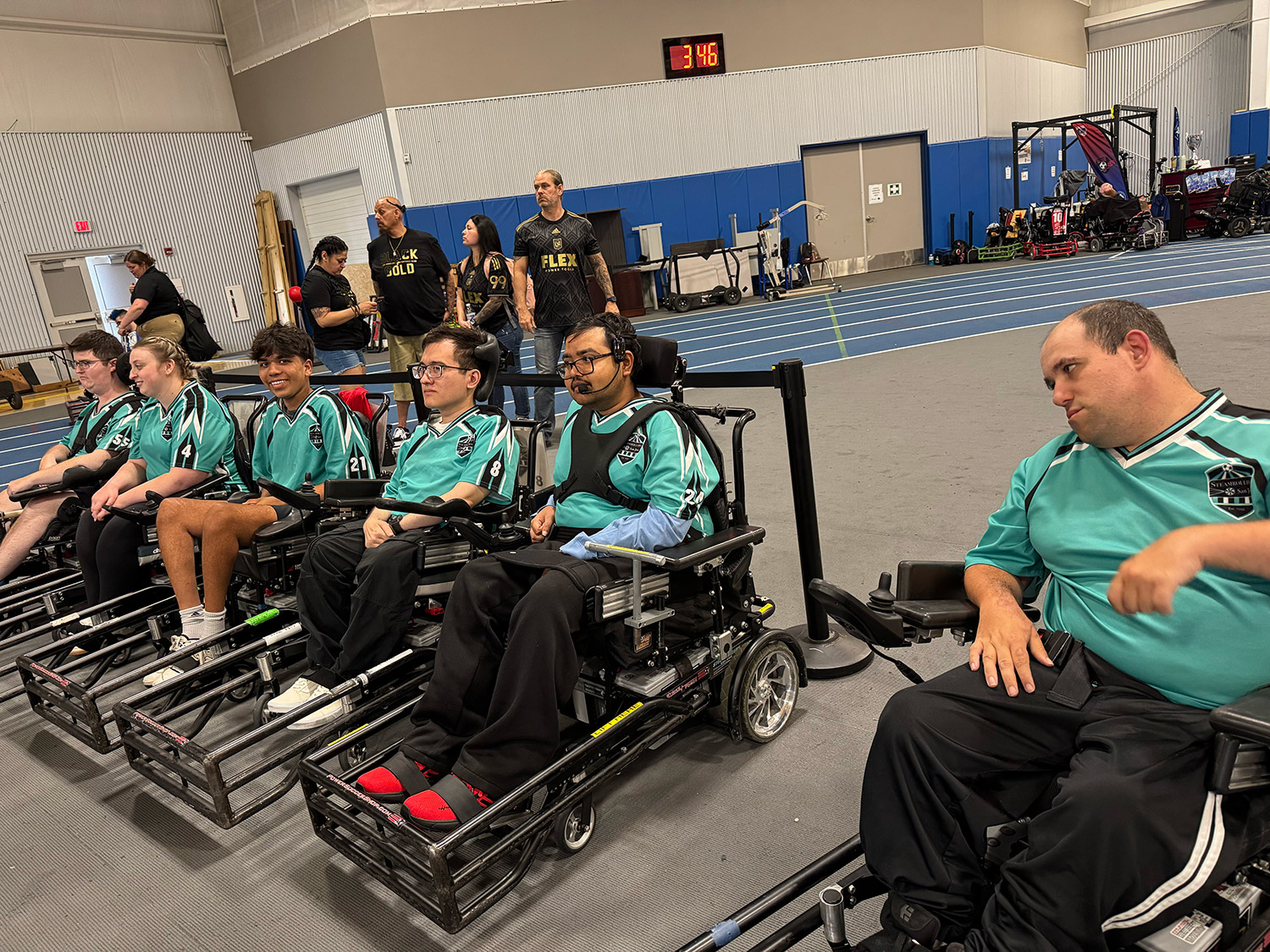A group of six people in teal sports jerseys sitting in power wheelchairs lined up indoors on a sports court.