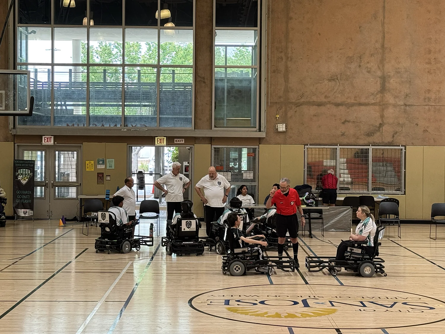 Indoor gym setting with power soccer players in wheelchairs and a referee in a red shirt talking to two players facing each other.