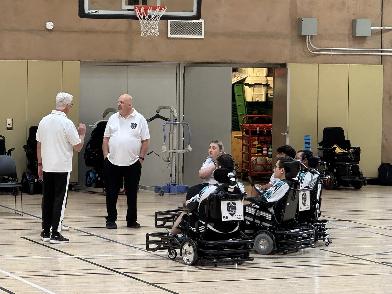 A group of people in powered wheelchairs and two standing men in white shirts are gathered on an indoor basketball court.