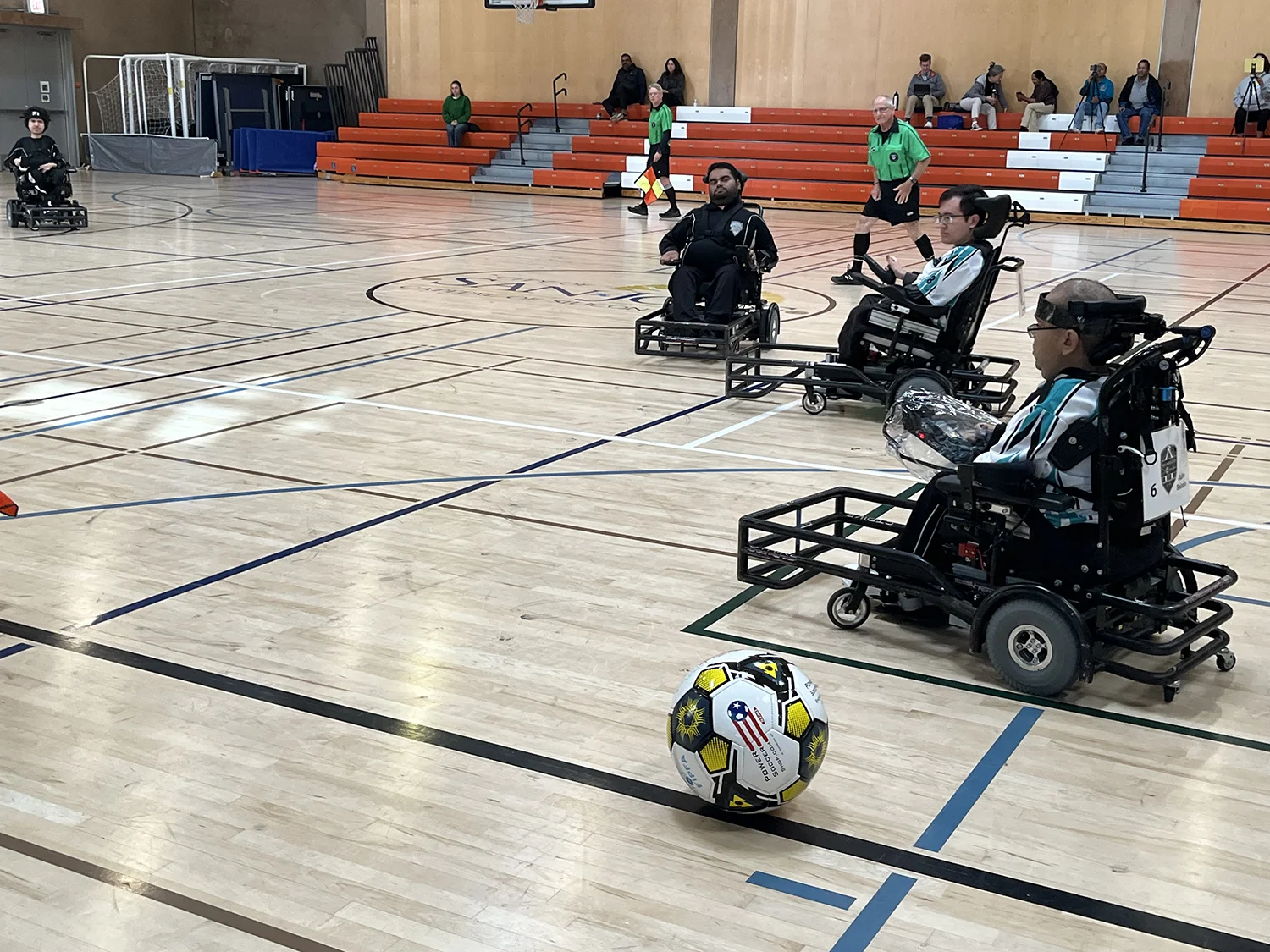 Players in motorized wheelchairs competing in an indoor power soccer game with a refereed gymnasium background.