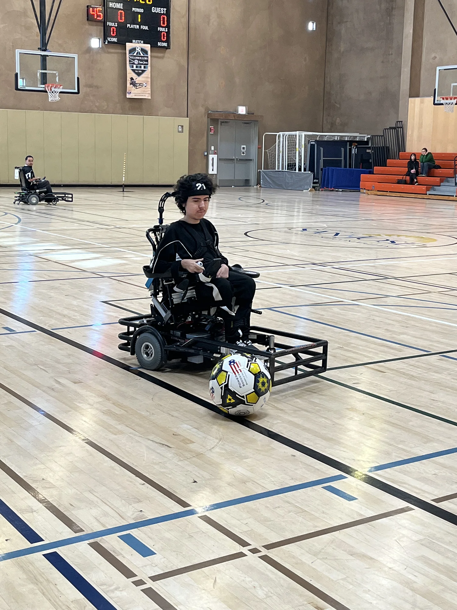 Two players in motorized wheelchairs playing power soccer on an indoor basketball court with scoreboard showing 45 seconds left in period one.