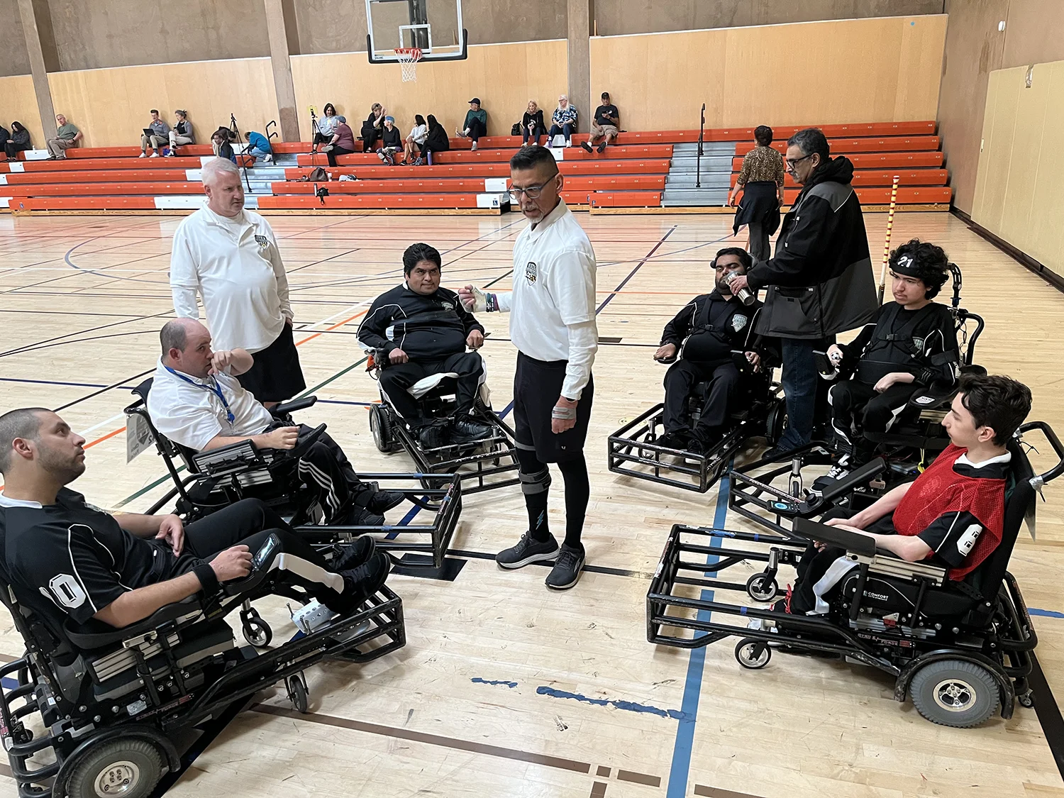 Group of athletes in power wheelchairs and coaches gathered on an indoor basketball court.