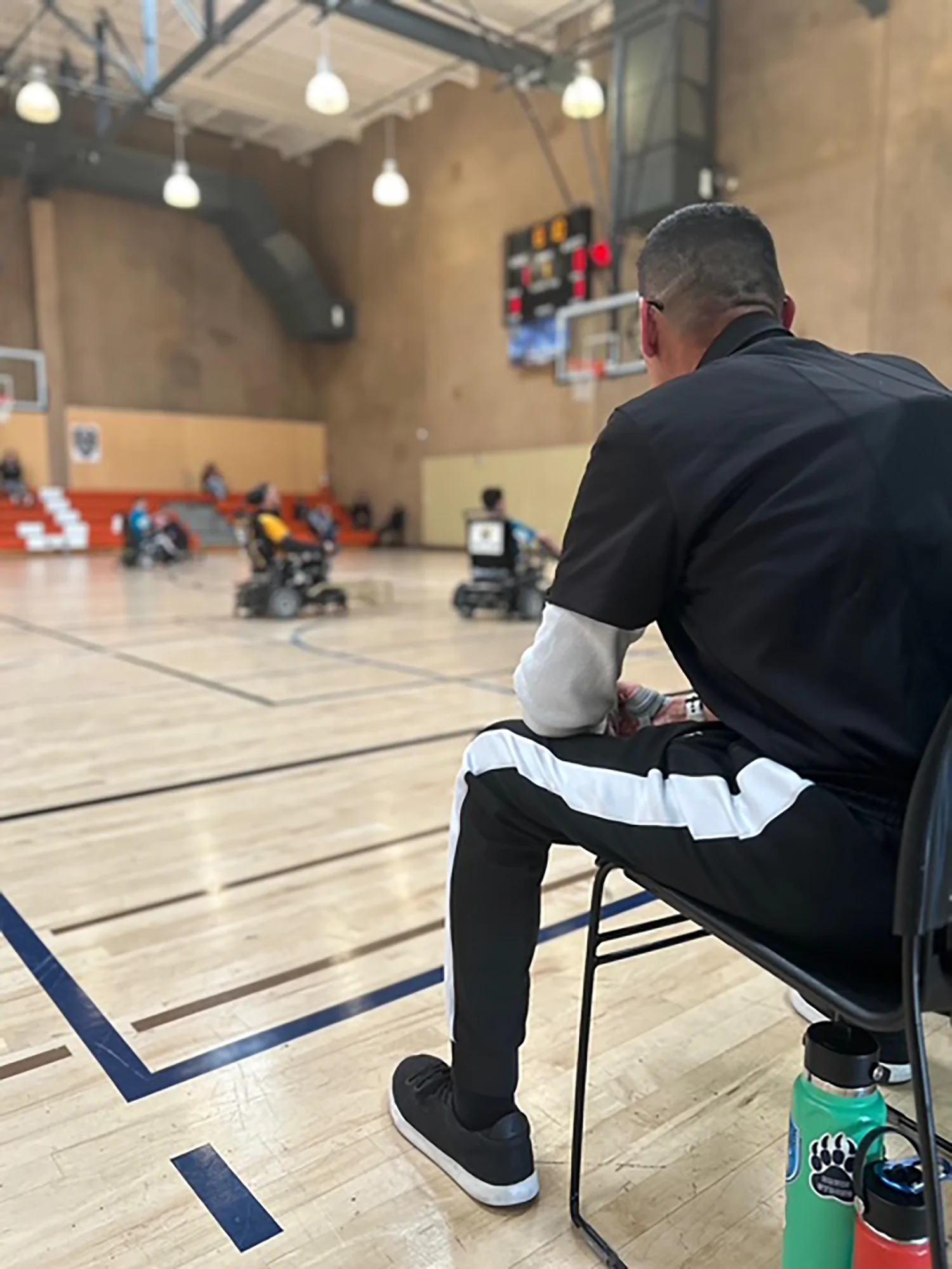 Man in black and white sportswear sitting on a chair watching a wheelchair basketball game in an indoor gym.
