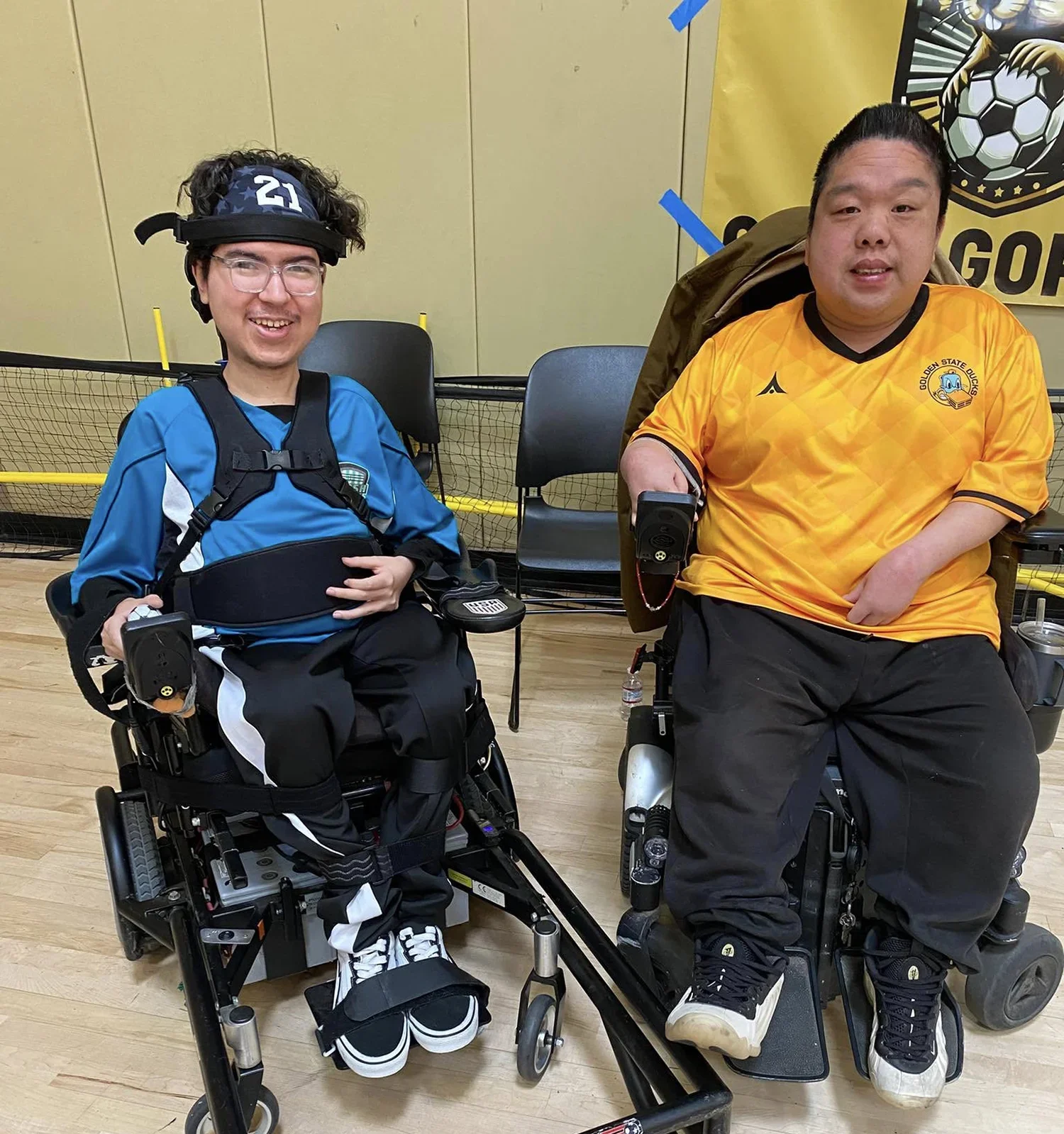 Two men in electric wheelchairs smiling and posing indoors on a wooden floor, one wearing a blue shirt and the other a yellow soccer jersey.