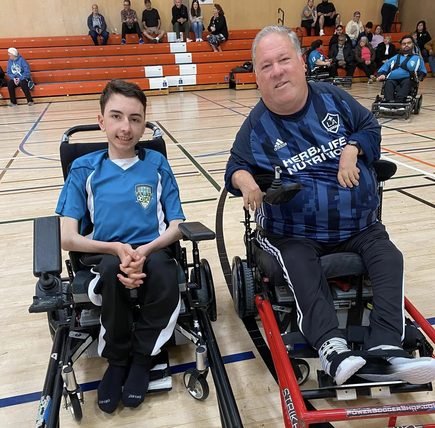 Two males in sports wheelchairs on a gymnasium floor wearing athletic jerseys, with bleachers and spectators in the background.