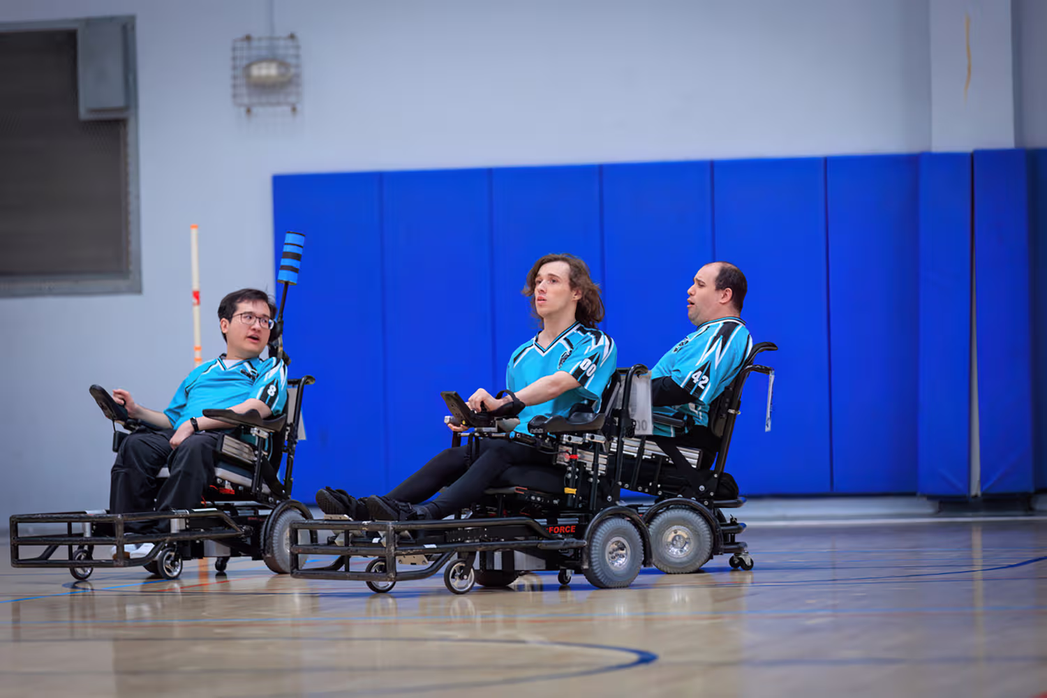 Three athletes in blue jerseys sitting in power wheelchairs on an indoor sports court.
