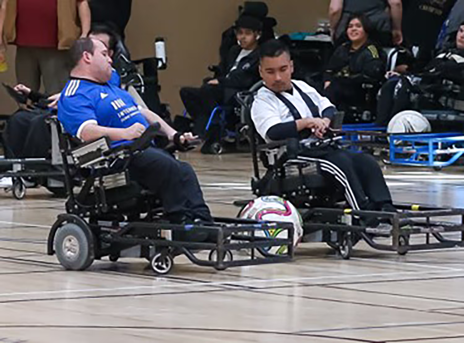 Two athletes in power wheelchairs competing in a power soccer match indoors with a soccer ball between them.