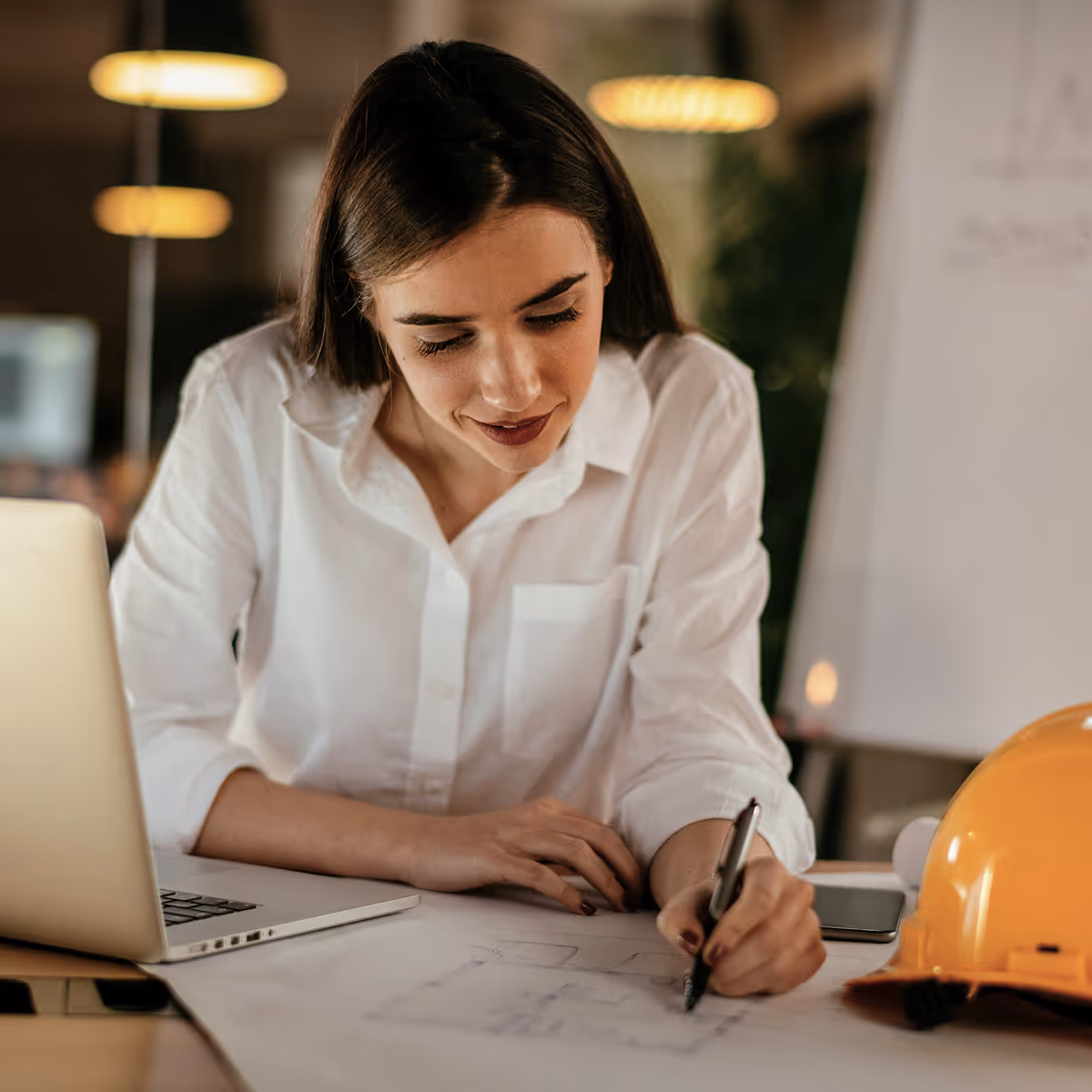 Construction site supervision architect over the plans and table with computer attentively looking through papers herself in white shirt.