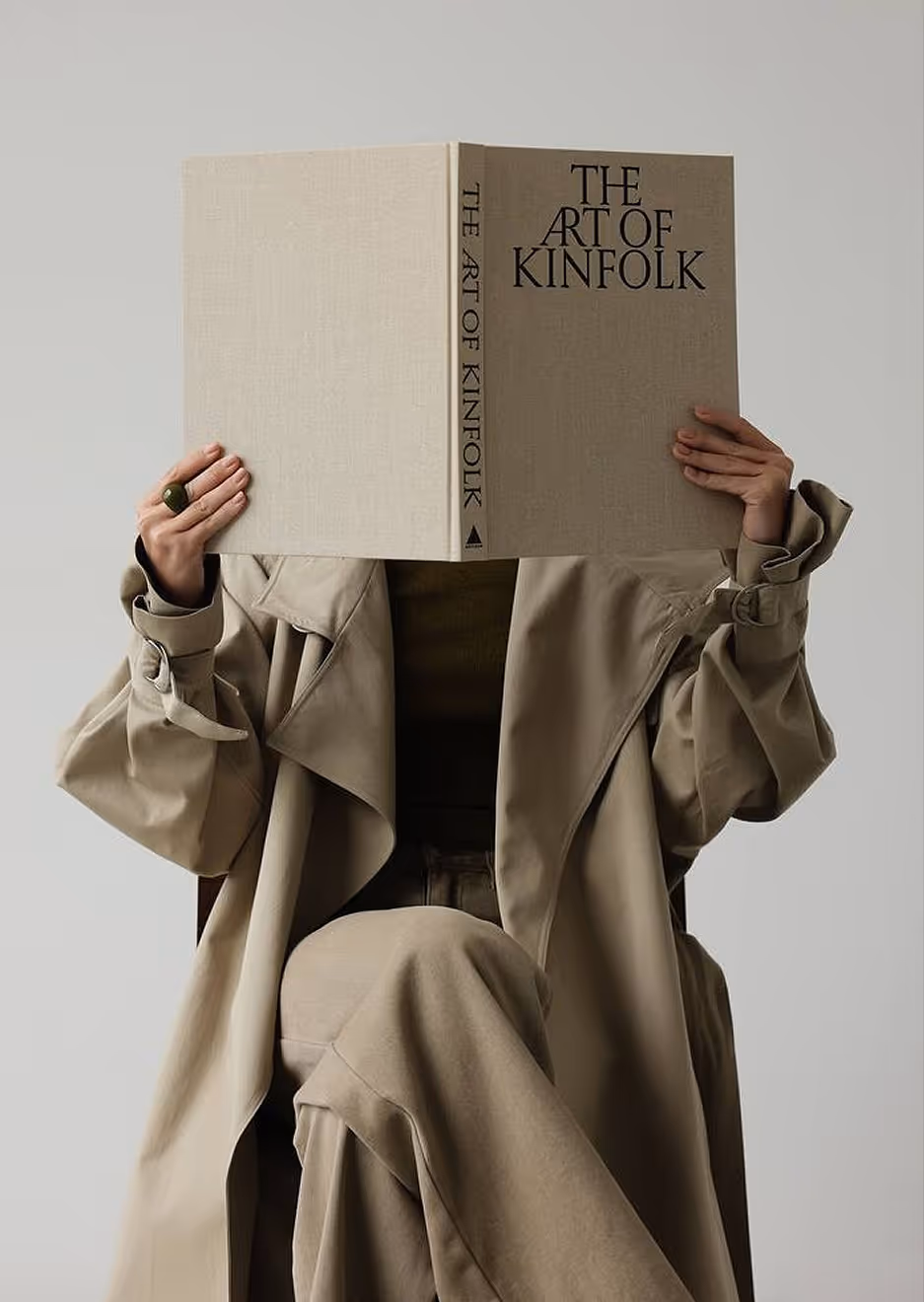 Lady in coat sitting on chair in bright space with a book covering her face deliberately in humor signature.