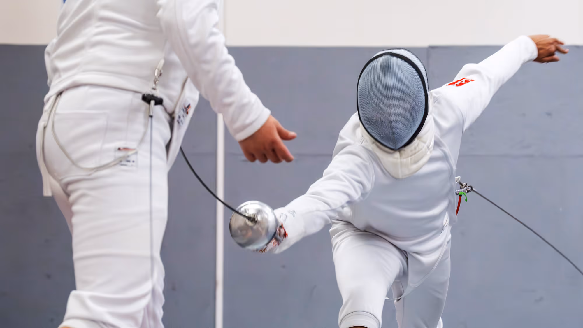 Two fencers in white gear engaged in a match, one lunging forward with a foil while the other prepares to defend.