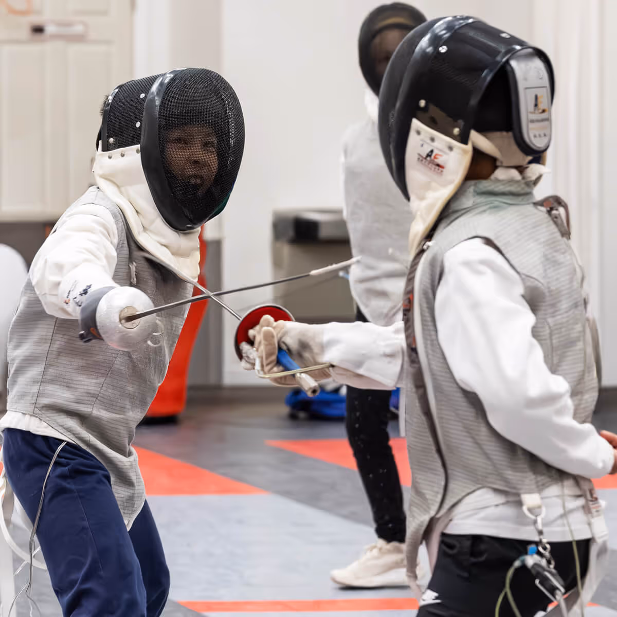 Two young fencers in protective gear facing each other with crossed swords during a match indoors.