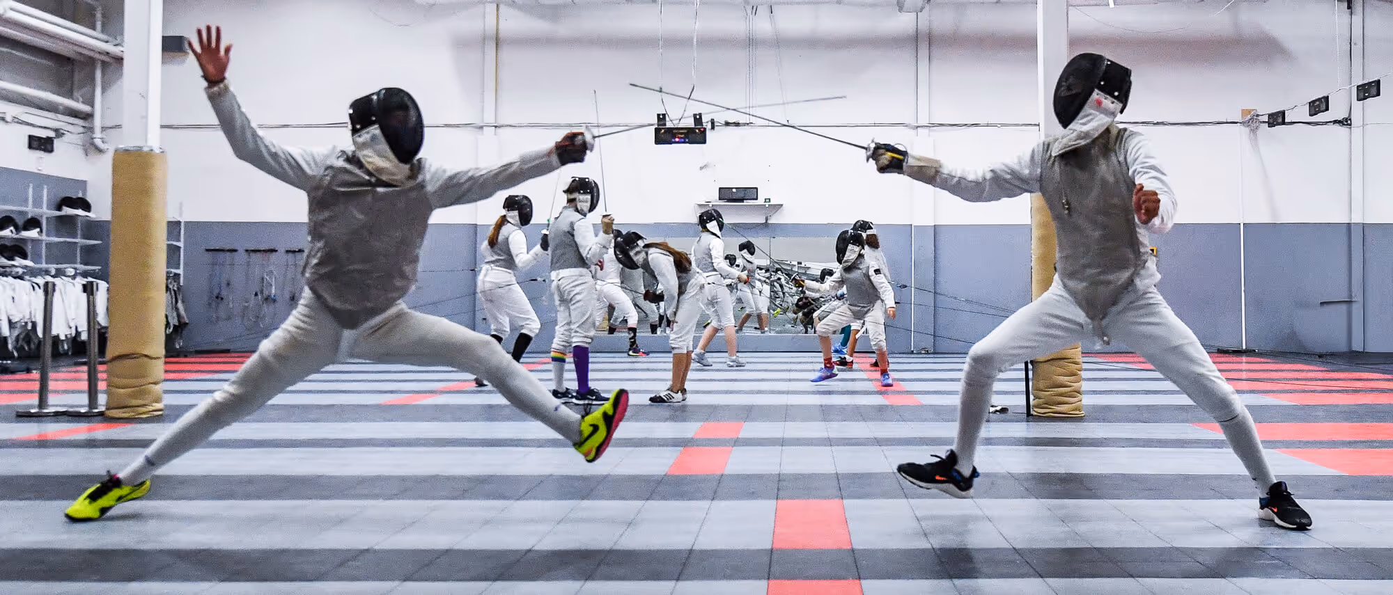Two fencers lunging toward each other in a fencing gym with other fencers practicing in the background.