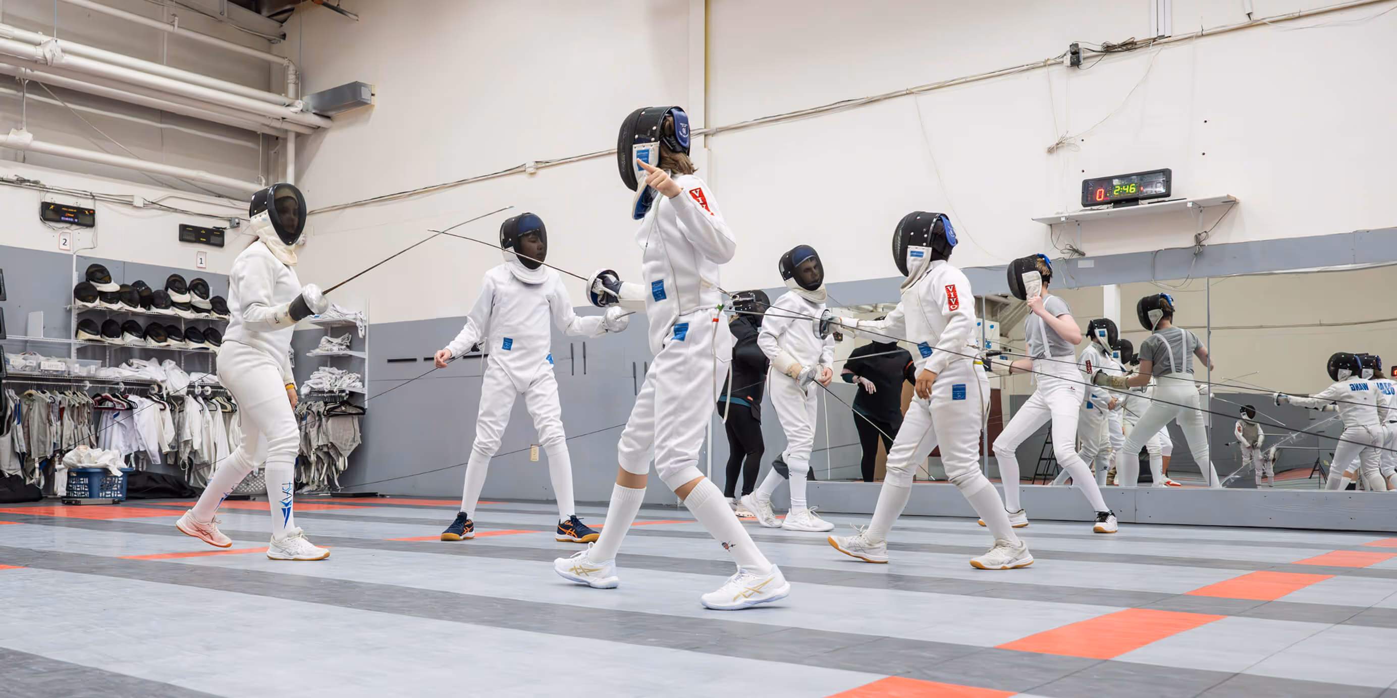 Group of fencers wearing white gear and protective masks practicing with swords in an indoor fencing training room.