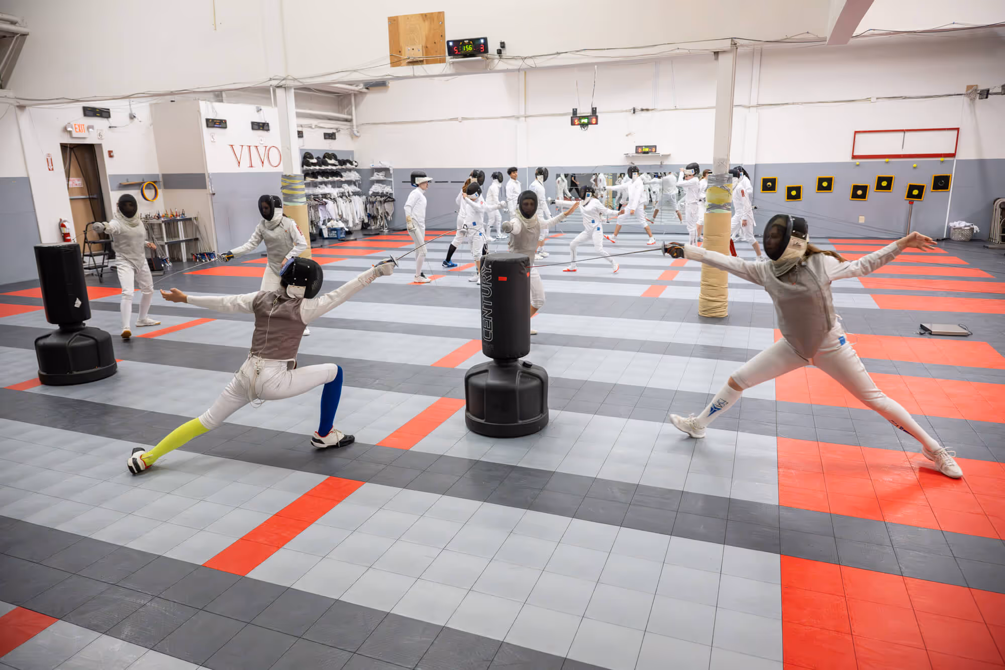 Fencers practicing lunges and sparring with foil swords in a large indoor fencing gym.
